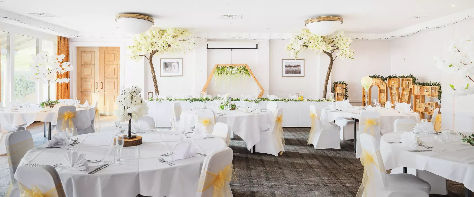 A large room set up for a wedding breakfast. Large tables are decorated with white flowers..
